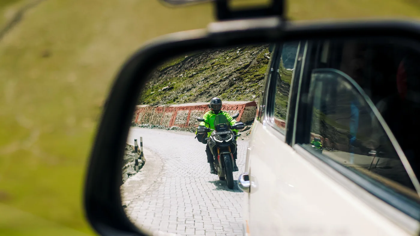 Motorcyclist in a high-visibility jacket seen in a car's side mirror on a winding road