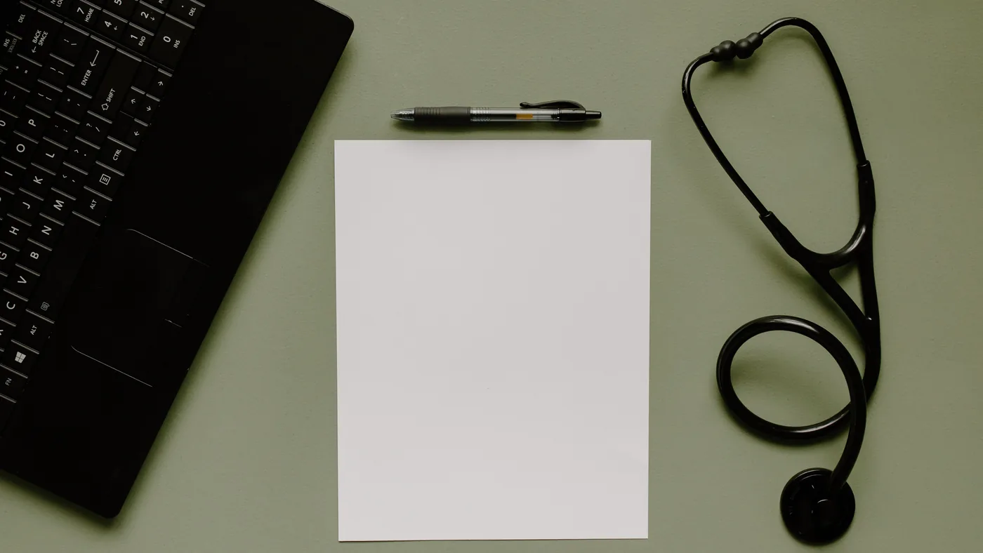 Stethoscope, a blank sheet of paper, a pen and a laptop on a desk, seen from above