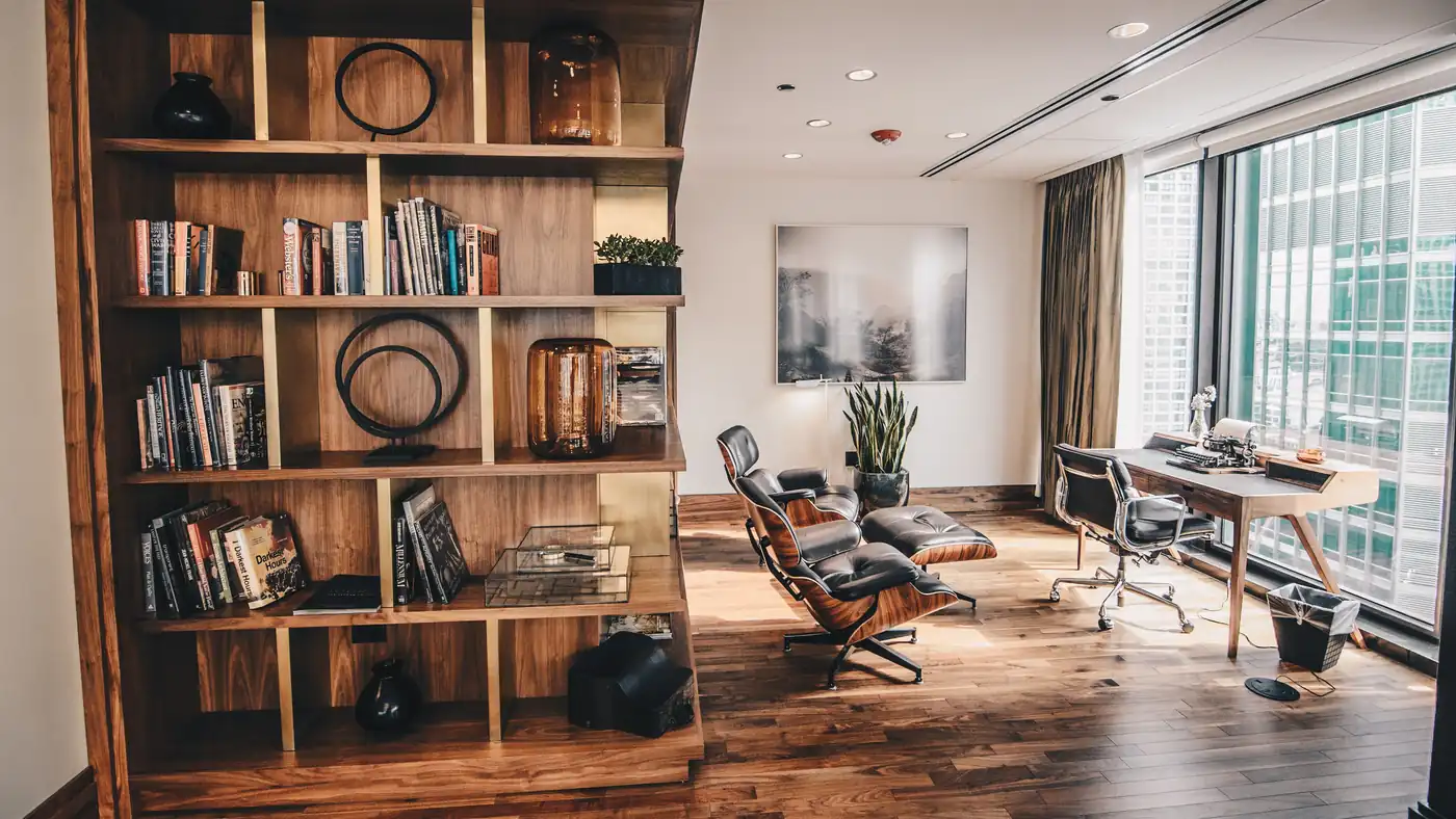 Office with a tall wooden bookshelf, a lounge chair and a desk by floor-to-ceiling windows