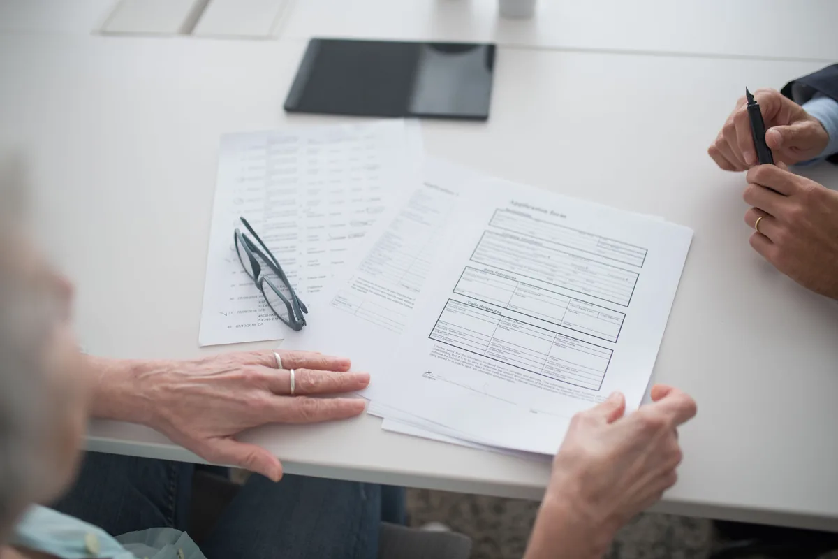 Two people reviewing printed forms at a desk beside a tablet and eyeglasses