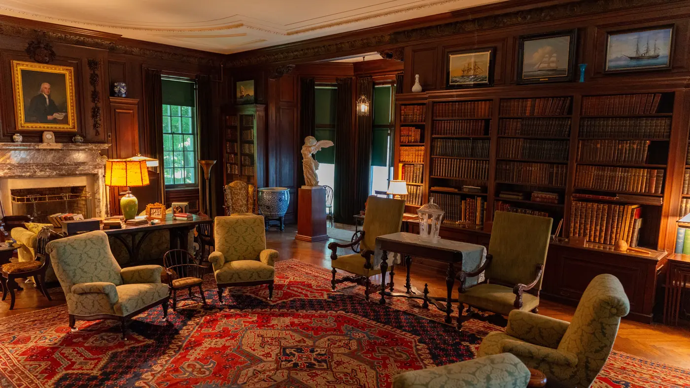 Wood-paneled library with leather-bound books on built-in shelves, armchairs and a red patterned rug
